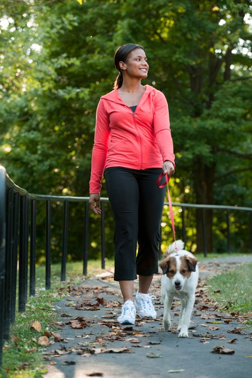 A photo of a woman walking a dog down a wooded path.