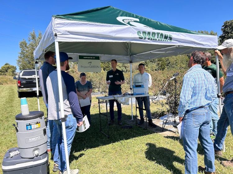 Several people stand under a canopy tent next to blueberry bushes during a field day.