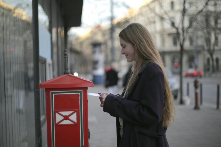 A white female with long blond hair wearing a dark coat sticking an envelope into a red mailbox on the street.
