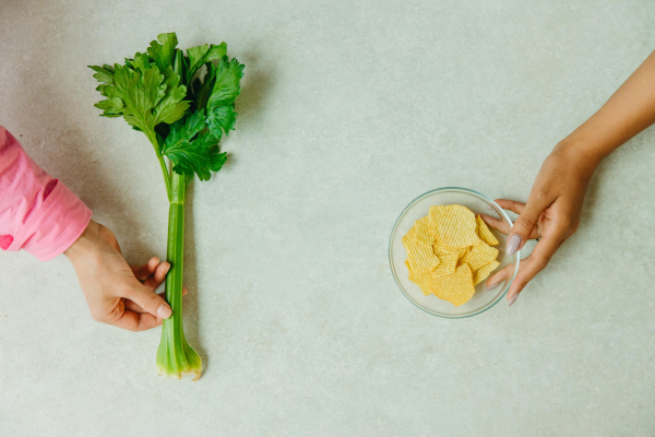 A photo of a hand holding a celery stalk and another hand holding a small bowl of chips