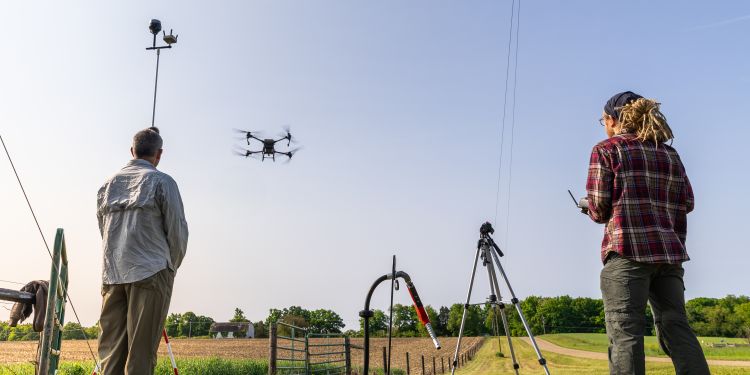 Two researchers stand at the edge of a field of crops while flying drones over the field.
