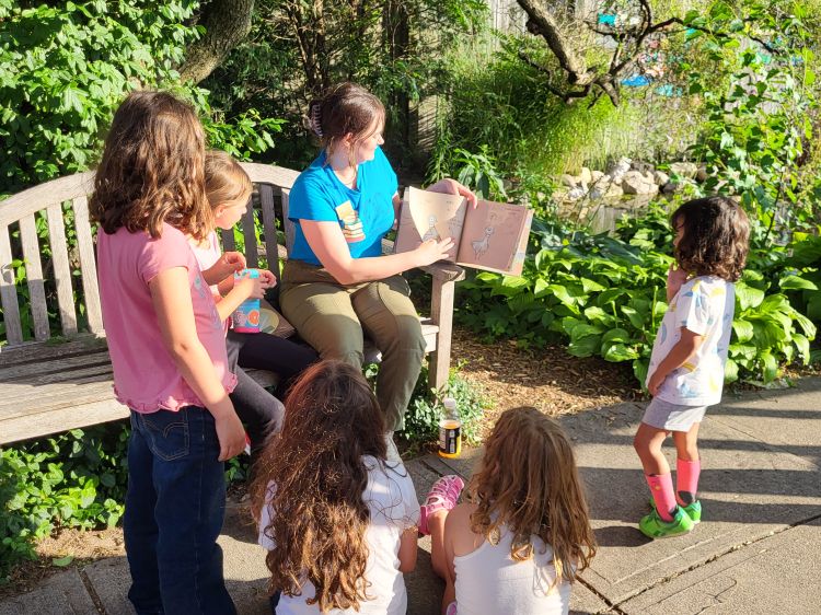 A woman sits on a bench in a garden reading a story to five children.