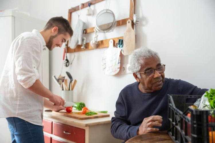 A man slicing tomatoes at a kitchen counter while an elderly person sits nearby.