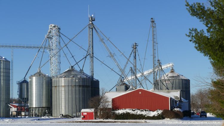 A snowy winter scene featuring several large metal grain bins and tall grain-handling structures under a clear blue sky.