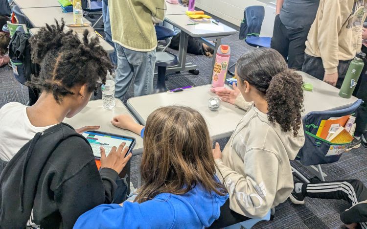Three youth at desks on a tablet moving a robot.