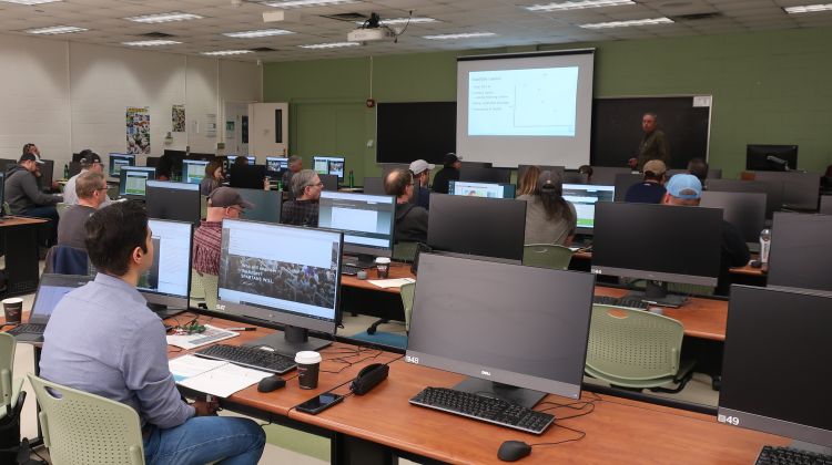A classroom filled with people sitting behind rows of tables with computers.