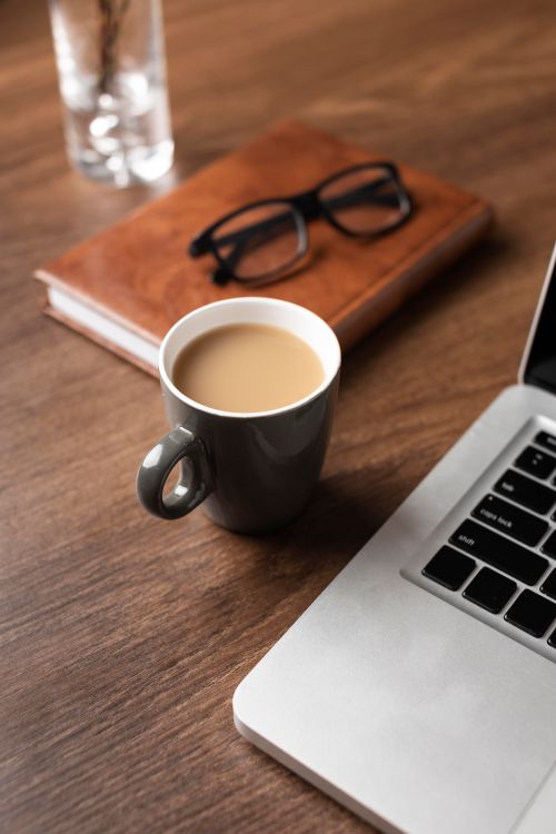 A coffee cup sitting next to a computer and a notebook with glasses.