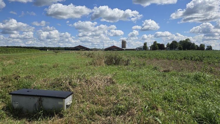 A farm landscape with green fields under a partly cloudy blue sky. In the foreground is a gray saturated buffer system box, with farm buildings, a silo, and a farmhouse visible in the distance.