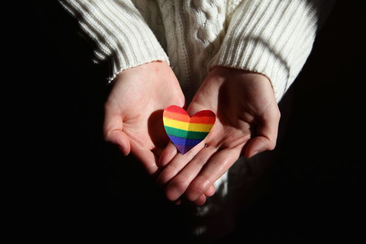 White hands outstretched holding a rainbow colored heart.
