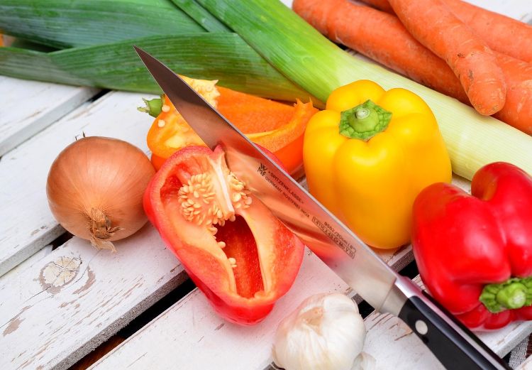 A photo of a knife cutting through a red pepper next to an assortment of vegetables.