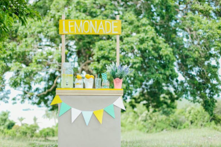 A grey and yellow simple stand in a green, outdoor area. A colorful banner and lemonade supplies adorn the stand.