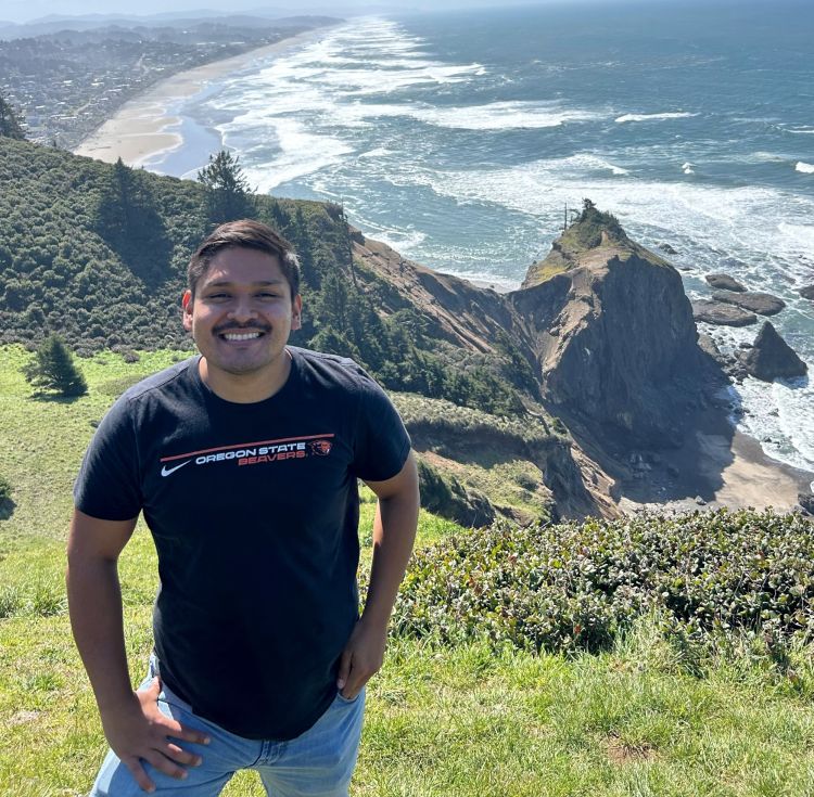 A smiling man in a black Oregon State Beavers t-shirt stands on a grassy cliff overlooking the Pacific Ocean, with rugged coastal bluffs, waves, and a distant shoreline in the background.