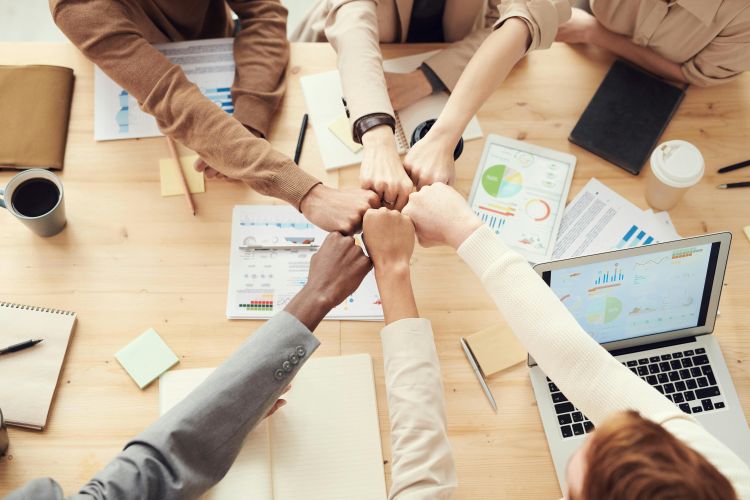 Six diverse hands coming in for a collective fist bump over a table filled with papers and laptops.