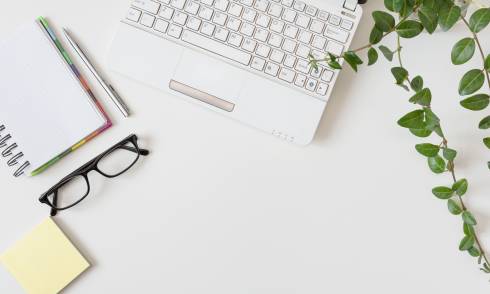 A while tabletop with note pad, glasses, laptop, and plant.