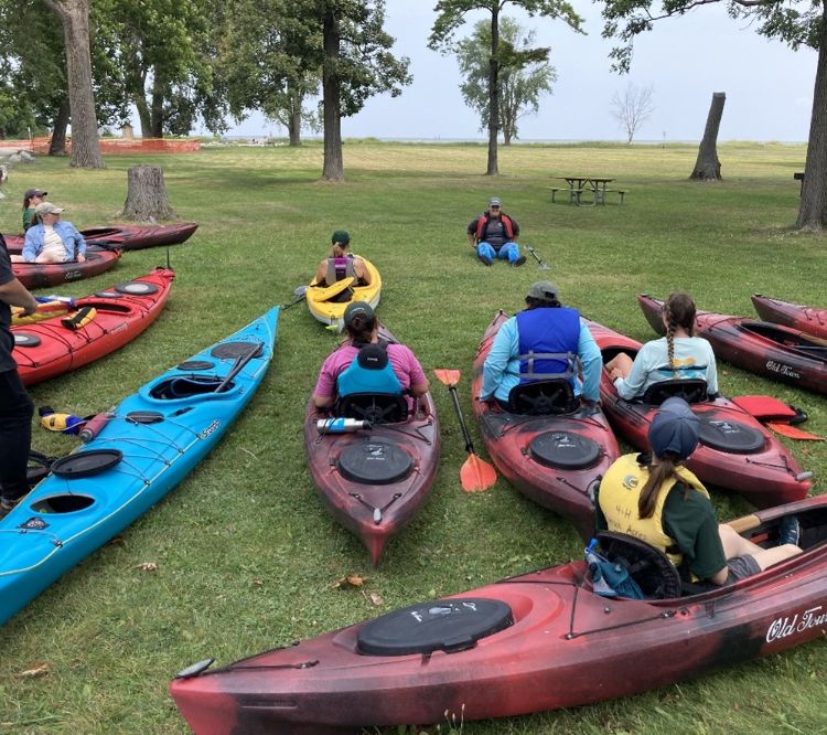 A group of people sitting in kayaks on the ground are being instructed in safety techniques.