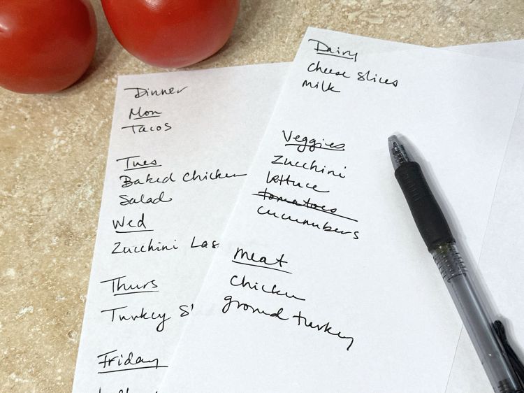 A hand written grocery list with a pen and some tomatoes on a countertop.