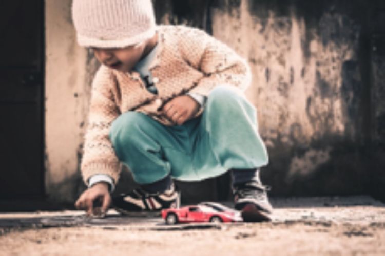 A young child with a hat on playing on the ground with a red car.