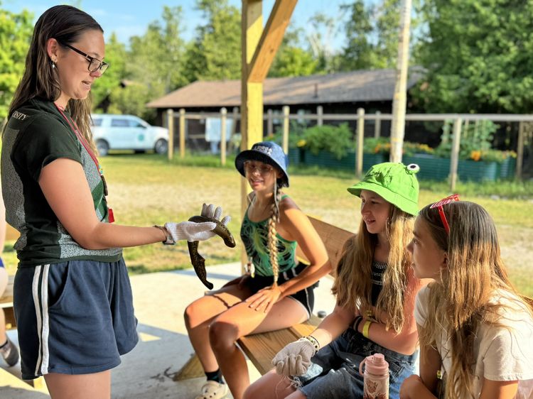 Woman standing to the left is holding a sea lamprey and displaying it to three teenagers sitting outside on a bench.