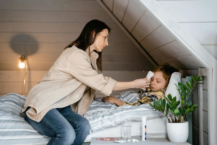 A parent takes a sick child's temperature while the child rests in bed.