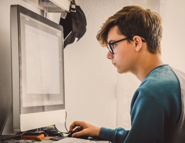 A young white boy in a blue shirt with dark hair and dark glasses sitting at a computer.