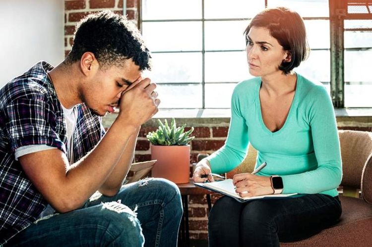 Two people having a discussion. The male has his hands over his forehead and is looking down. The woman is looking on intently taking notes.