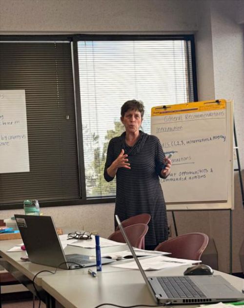 A woman speaks at a meeting in a conference room with a large easel notepad behind her.