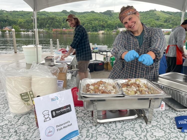 Two people stand inside a food prep area cooking whitefish for visitors to taste.