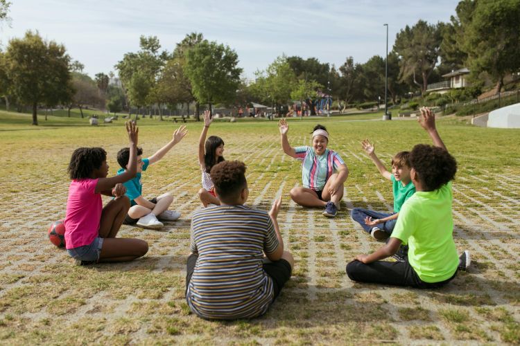 Diverse sitting in a circle in a field with raised hands.