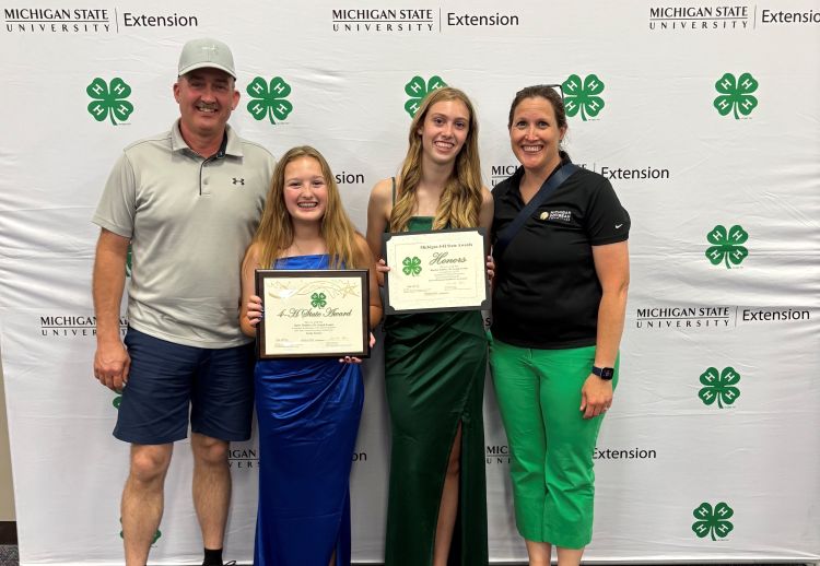 A husband and wife with their two daughters holding up their awards in front of the MSU Extension Michigan 4-H backdrop.