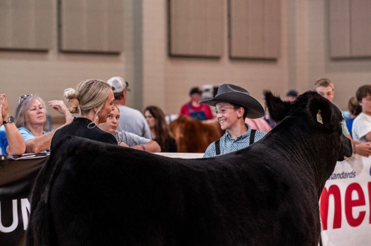 A young man in a cowboy hat leading a black calf shaking the hand of a woman in the ring.