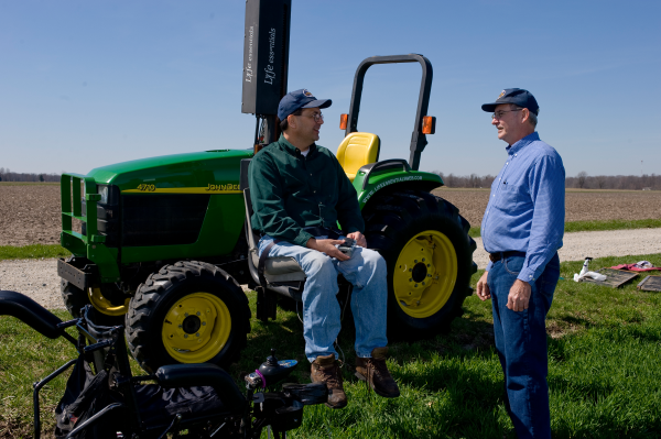 Two men talking; one is standing and the other sits on a lift mounted on the side of a green tractor with rollover protection bars.