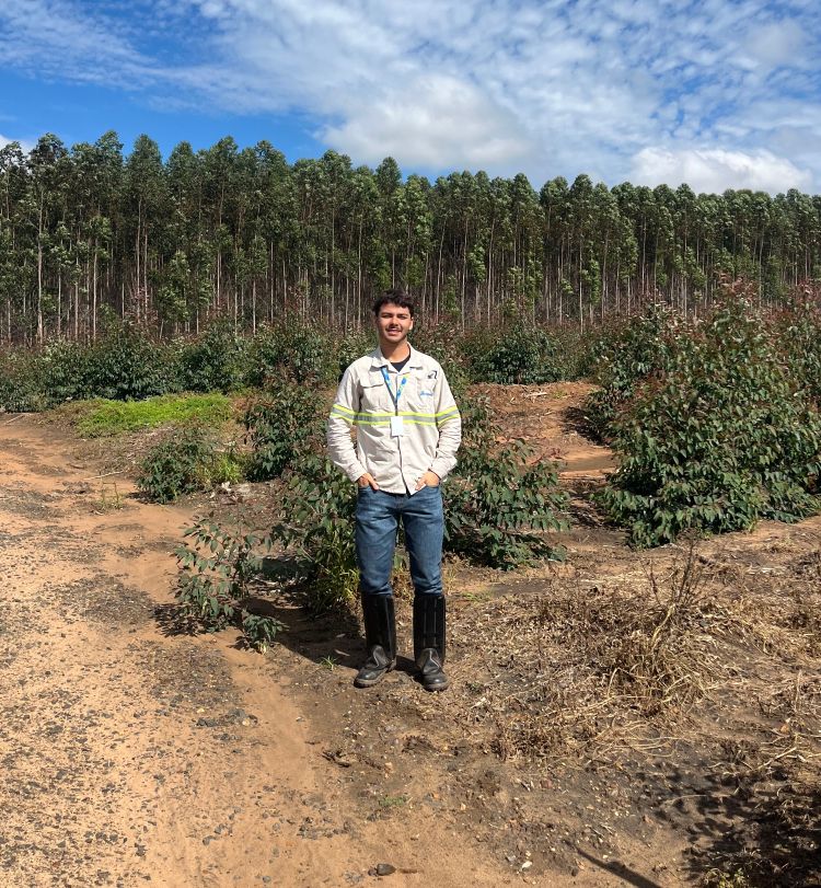 A forestry student standing in a field with forest trees in the background.