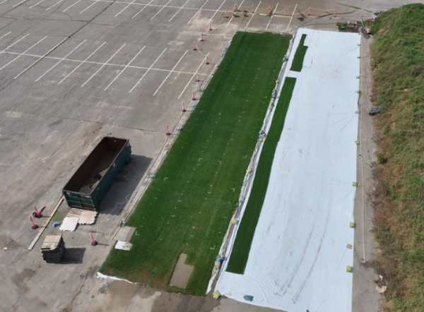 Aerial image of a strip of sod in a parking lot, there are parking spaces to the left and a strip of white material to the right. To the far left there is a large dumpster.