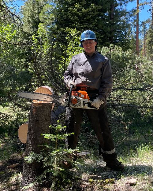 Riley Watts, MSU Forestry student posing for a picture holding a chainsaw with a tree cut down next to them.