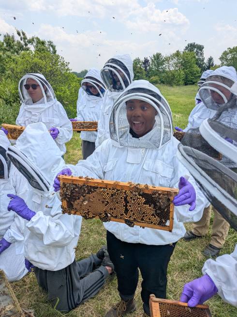 A woman smiles while holding a honey frame in a bee suit, surrounded by other beekeepers.