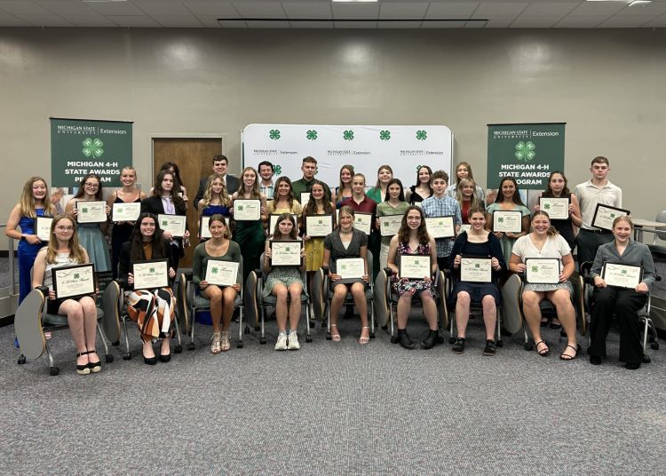 A group of young people sitting and standing in front of a 4-H banner with plaques in their hands.