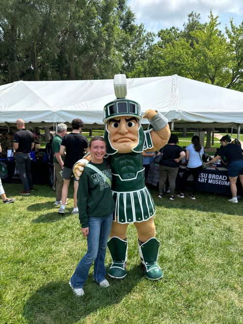 A young lady in blue jeans and a green MSU sweatshirt standing next to the Sparty mascot.