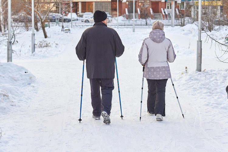 A photo of two older adults walking through the snow using trekking poles.