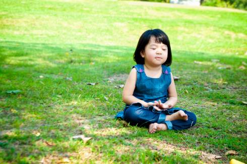Image displays a little girl sitting in the grass, hands together and legs crossed.
