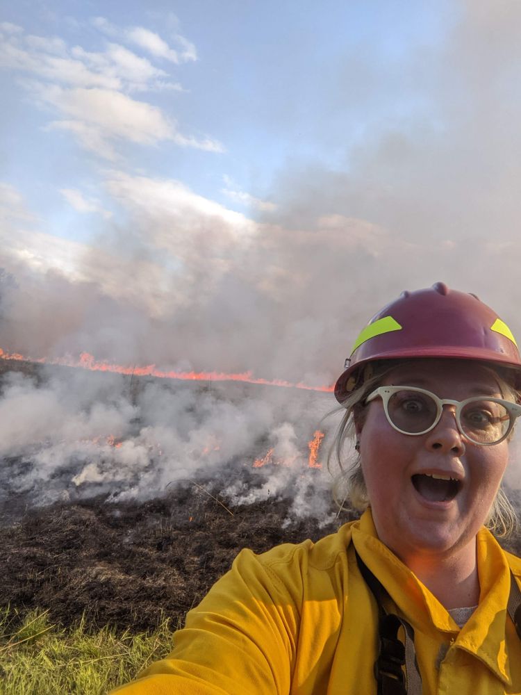 Erin Budzyn, PhD Candidate, posing for a selfie in front of a field with fire.