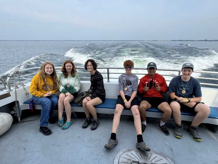 Six students sit on a bench that stretches along the back of the Lady Michigan boat. The wake of the boat is seen in Lake Huron behind the students.
