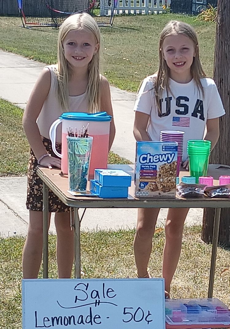 Two young blond girls standing behind a table with lemonade.