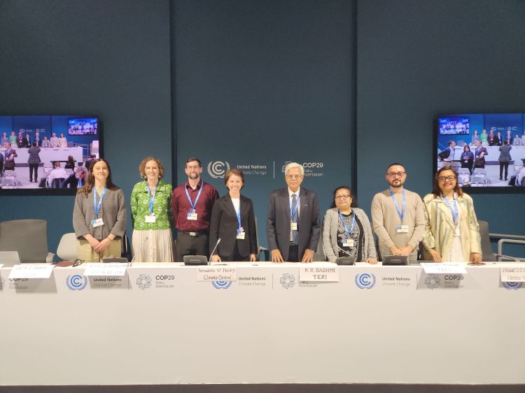 Eight people stand beside each other, smiling, behind a white table in a large meeting room for the COP29 meeting in Baku, Azerbaijan.