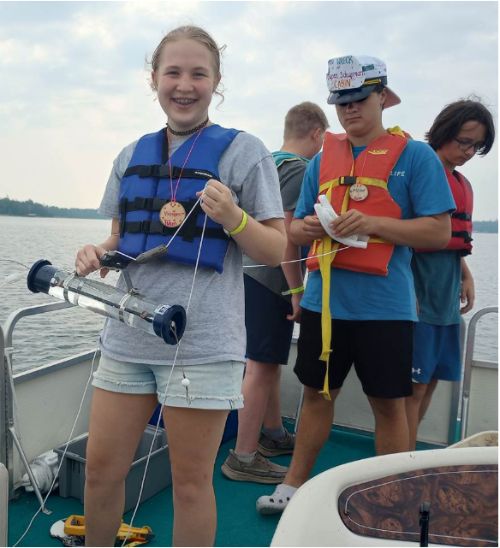 A young girl and boy on a boat wearing life jackets holding a water sample.