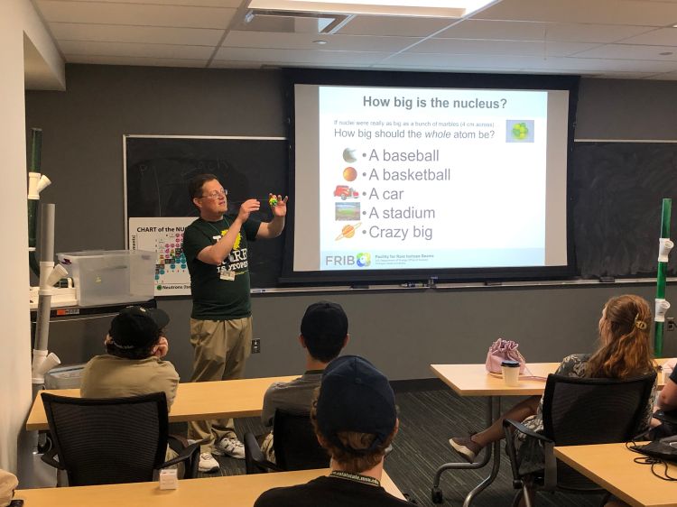 Youth in a classroom setting facing a man standing in front of the room standing next to a projected image, gesturing.