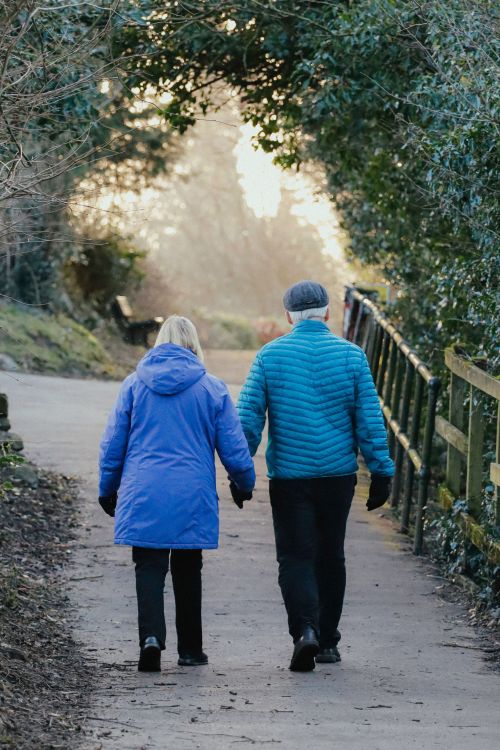 Two older adults holding hands and walking down a park pathway, it appears cold outside.