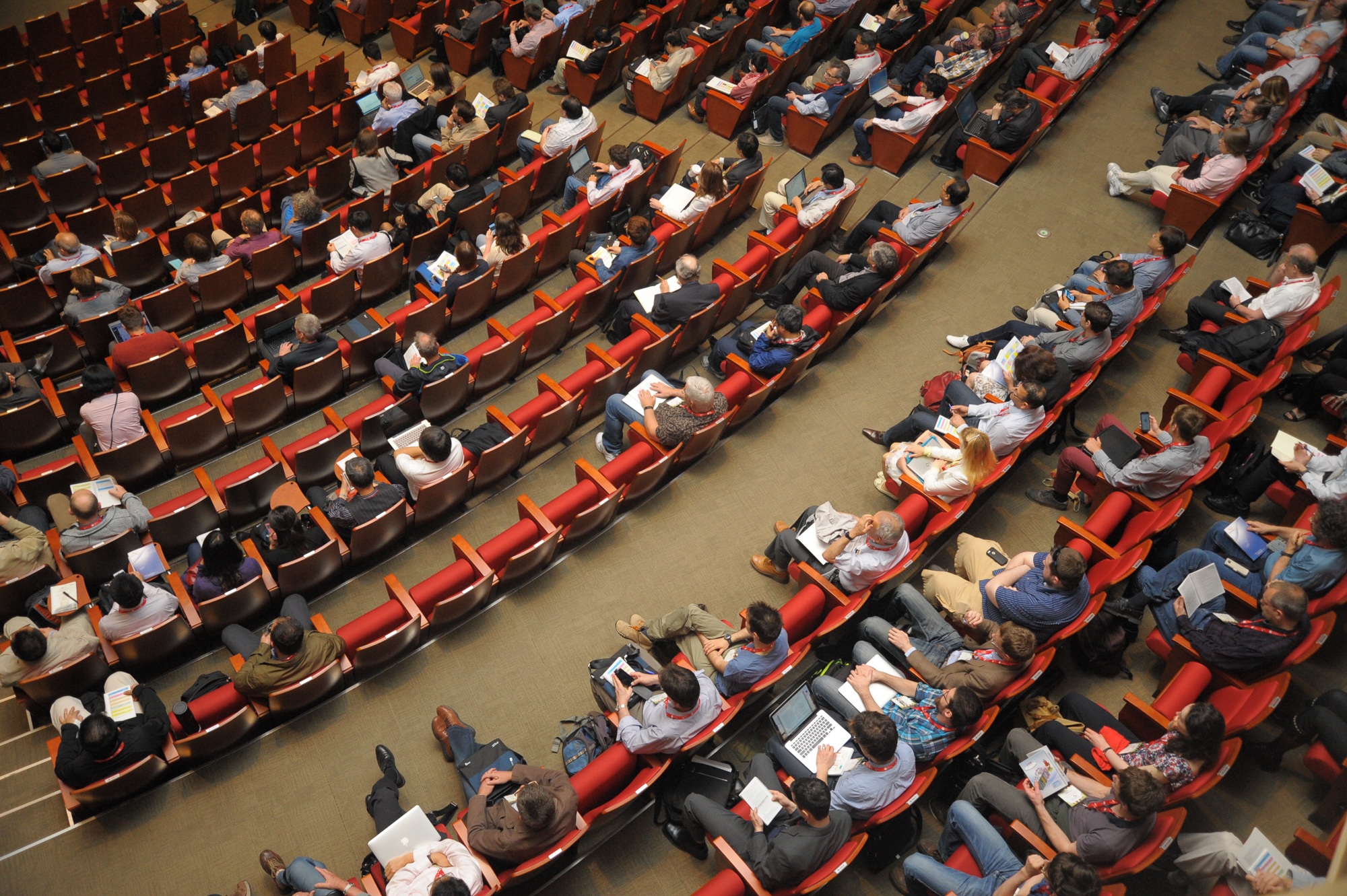 A large auditorium filled with conference attendees seated in curved rows of red chairs, viewed from above, with many participants holding laptops, notebooks, and printed materials.