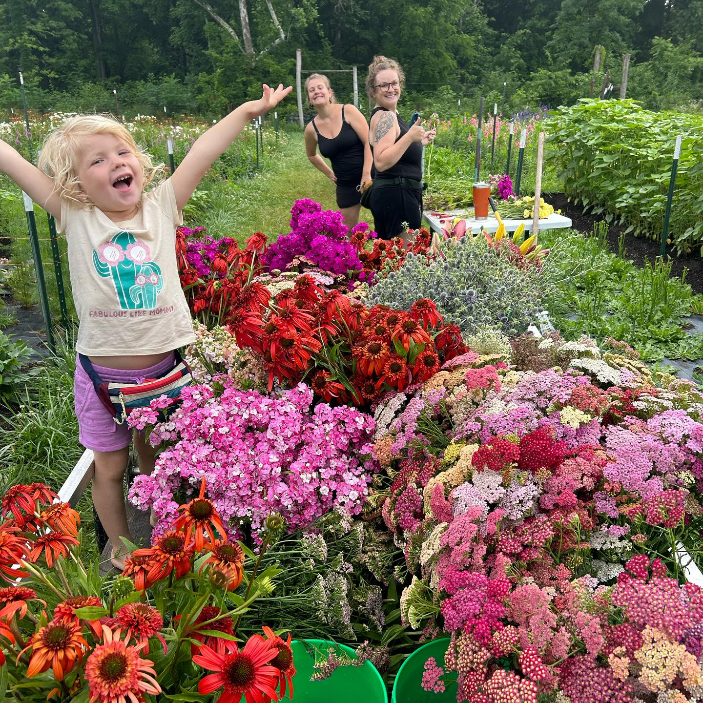 A row of colorful flowers with a young girl jumping in the air and two women in the back, cutting flowers.