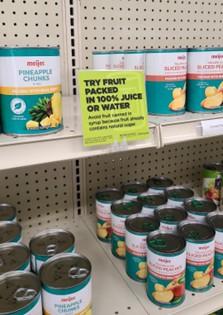 Cans of fruits on a supermarket shelf with a sign suggesting fruit in 100% juice or water.
