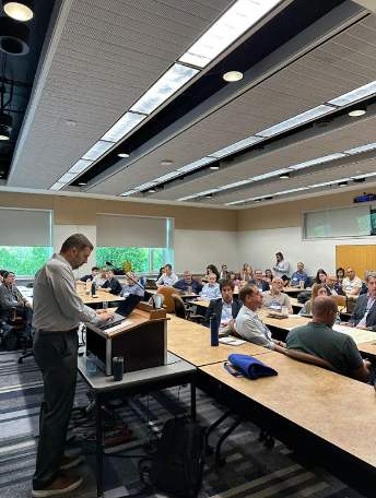 Photograph of a professional conference room with a speaker presenting at a podium to an audience seated at long tables arranged in rows. 
